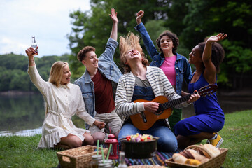Group of friends having fun on picnic near a lake, sitting on blanket eating and playing guitar.
