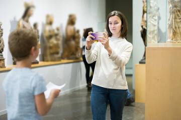 Portrait of young woman using phone in museum, photographing school age son near exhibition