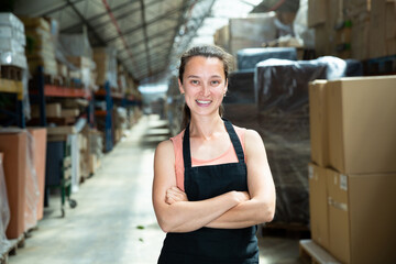 Positive female in apron standing near shelves with cardboard boxes for packaging in the stock