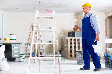 Man painter in coverall and hardhat carrying bucket and paint roller. Male builder working in...