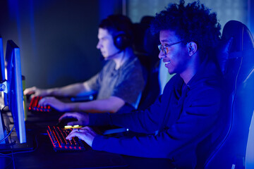 Fototapeta na wymiar Side view portrait of smiling African American man playing video games in cybersports club in blue neon lighting