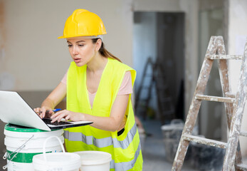 Female foreman in a protective helmet and a yellow vest checks the execution of repair work using a...