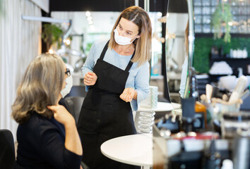 Female hairdresser wearing protective mask to prevent spread of viral infections during pandemic offering hairstyling to woman client visiting professional hair salon