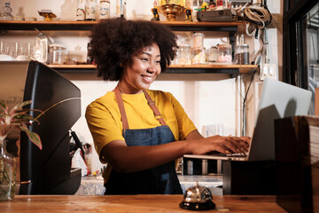 African American female cafe barista in casual apron happy with online customer order in laptop, cheerful works with smile at counter bar in coffee shop, small business startup entrepreneur lifestyle.