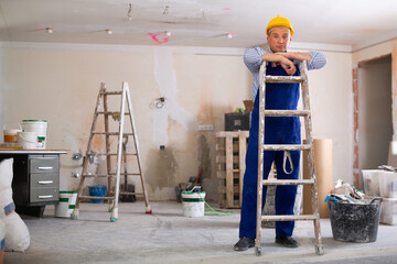 Worker in blue overalls carries a ladder on his shoulder in a room being renovated