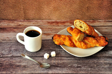 Two spiral puff pastries on a plate, a cup of black coffee on a wooden table, a spoon and two sugar cubes nearby. Close-up.