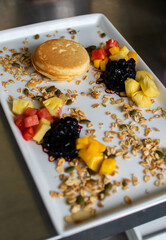 Homemade pancakes with granola, honey, watermelon slice, pineapple, berries, and whipping cream on white plate and aesthetic textured background