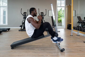 Young short curly black hair man with moustache and beard Sit-up on Multi-Function Bench. There are cardio machines and dumbbell set with rack in the gym.