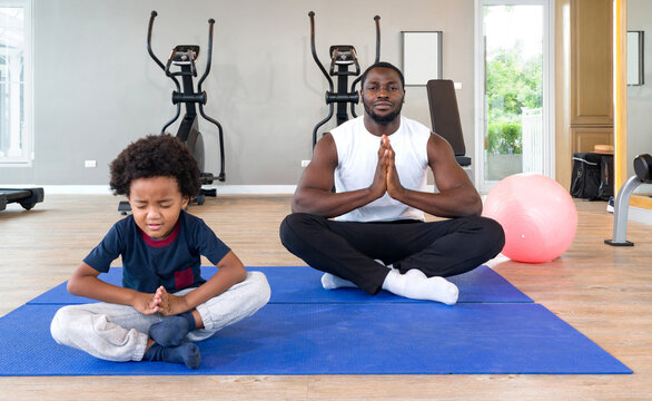 Young short curly black hair father  with moustache and beard meditating on yoga mat before exercise. The boy sit unhappy in the front. Morning fitness, mindfulness concept. - Powered by Adobe