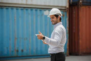 Engineer worker in causual suit standing in shipping container yard holding laptop with smile. Import and export product. Manufacturing transportation and global business concept