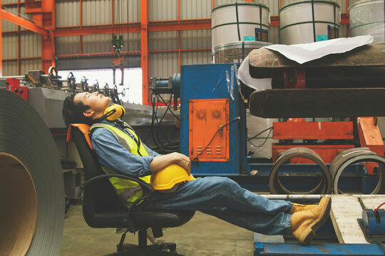 Asian male worker takes off his helmet and sleeps stretched chair taking daytime nap during his break : Factory worker working hard tired male worker resting work area.