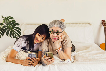 Portrait healthy mother and cute Asian daughter lying on the bed playing smartphone in the afternoon at home showing warm love to each other : Daughter always lies on her mother's shoulder with love.