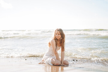 Fototapeta na wymiar Young blonde beautiful woman with long hair in white dress enjoying life on sea beach