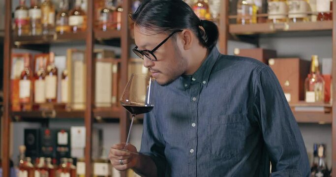 Young Asian sommelier sniffing and tasting newly imported wine and smiling happily after tasting. man wearing glasses and hair tie Wine tasting at the restaurant bar at the hotel.