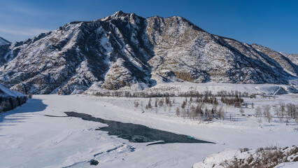 A frozen river in a snowy valley. The central part of the riverbed is not frozen. Steam over turquoise water. Bare trees on the banks. A picturesque mountain range against the blue sky. Altai. Katun
