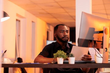 Executive manager analyzing paperwork report and taking notes to create research presentation in office with big windows. Working on project data with documents and computer during sunset.