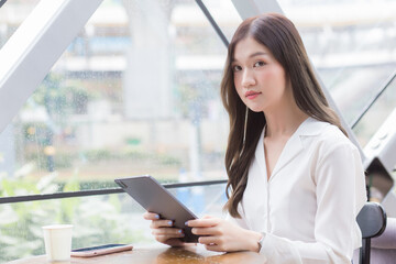 Obraz premium Professional beautiful young Asian business woman is looking at camera in her hands tablet or notepad while she is sitting at coffee shop among business background in the city.