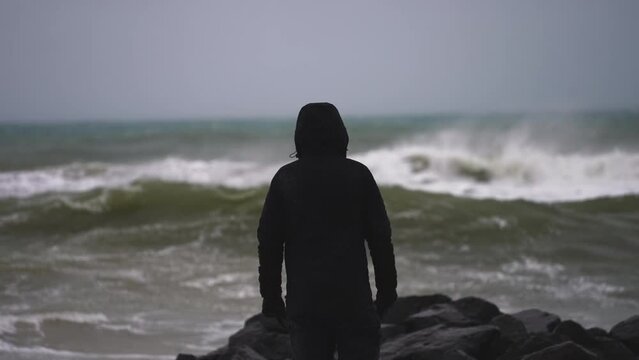 Man Stands by the Sea on a Rocky Coast and Watching Coming Storm and Big Waves