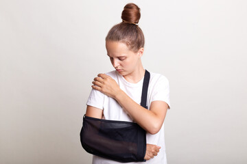 Indoor shot of unhealthy woman wearing casual T-shirt standing in arm sling and feeling pain in...