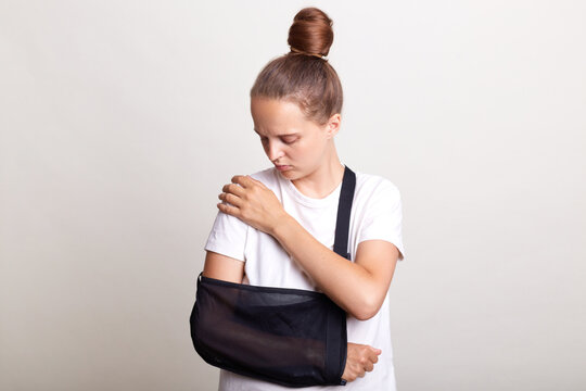 Indoor shot of unhealthy woman wearing casual T-shirt standing in arm sling and feeling pain in shoulder, touching painful part of the body, car accident, posing isolated on light gray background.