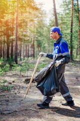 Man in overalls collects garbage and waste in forest. Ecological problem of pollution of nature. Young guy with plastic bag cleans forest on summer day.