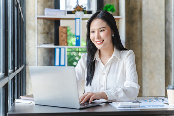 Fototapeta premium Young beautiful Asian woman in white shirt typing on computer laptop while sitting at the working wooden table over modern office as background.