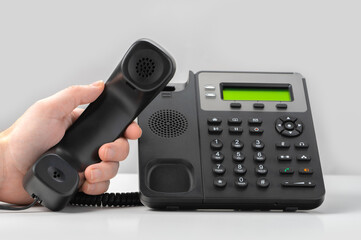 hand holding voip telephone receiver on gray background. black office landline voip phone with handset up on table.