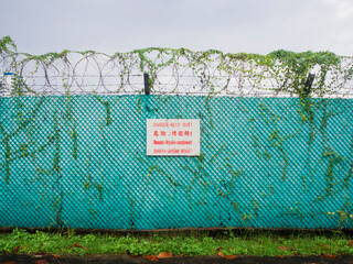 A "Danger-Keep Out!" sign in English, Chinese, Tamil and Malay on a barb wired green fence surrounding a restricted area. Plants creeping on the fence of an abandoned restricted property.