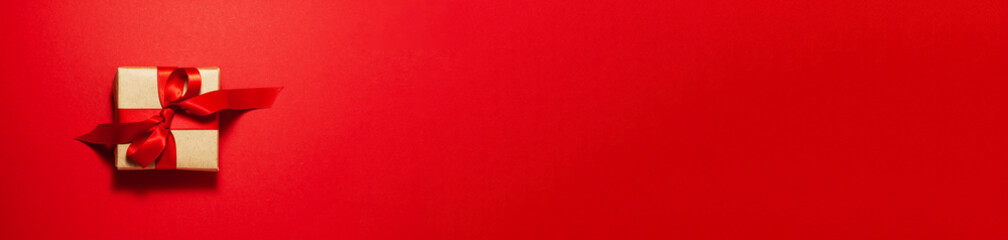 Banner of brown paper gift box with a red satin ribbon bow on top on red. Christmas holiday mother and father Valentines days and birthday present flat lay concept with copy space