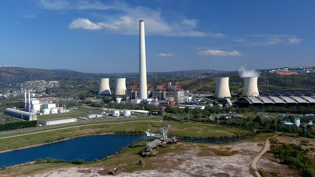 Thermal power plant with smoking chimney behind the lake and old drilling machine, bottom wind turbines on a sunny afternoon. Drone firing traveling forward. As Pontes, Lugo, Spain