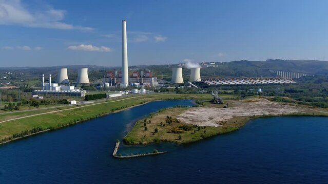 Thermal power plant with its smoking chimney, a lake in front with an old industrial machine, a bridge on the right, bright and sunny afternoon. Drone shot rising. As Pontes, Lugo, Spain