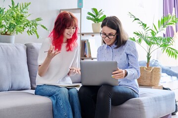Female teacher teaches teenage girl, sitting together on couch in office, using laptop