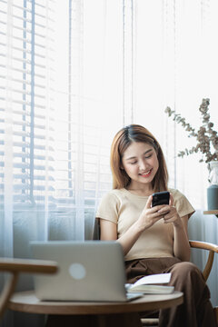 Happy smiling Asian woman relaxing using smartphone to talk and laptop computer in bedroom at home. creative girl working and typing on keyboard Study and work from home vertical image concept