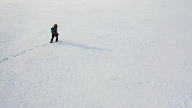 Lonely person in winter clothes with backpack and ski poles walks through endless snow-covered area, side and back view. Camera follows the hiker, the birds eye view.