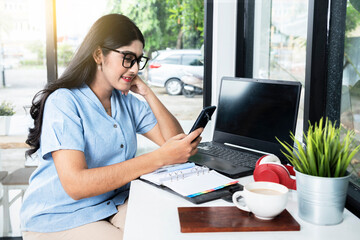 Asian woman with eyeglass using a mobile phone with a laptop and a notebook with a cup of coffee on the table