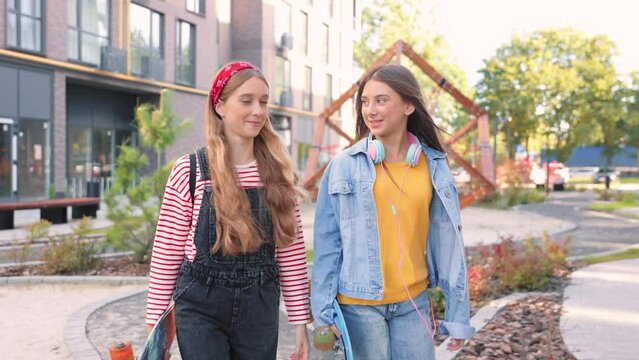 Portrait of Caucasian joyful young beautiful girls with skateboards walking on street and talking in good mood. Casual teens friends skateboarders outdoor in city, leisure, friendship, teenage concept