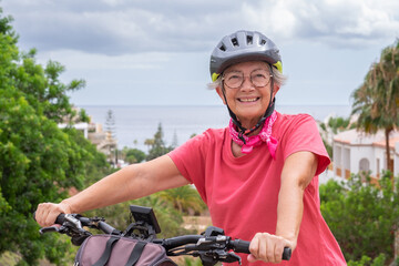 Cheerful elderly cyclist woman in pink jersey and helmet pushing her electric bicycle up the climb, sea behind, enjoying freedom and vacation