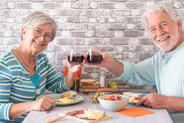 Smiling caucasian senior couple toasting with red wineglass while sitting face to face at table having brunch together at home