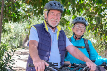 Naklejka na ściany i meble Portrait of happy active caucasian senior couple with electric bicycles running outdoors in the park. Two smiling seniors wearing cycling helmet enjoying a healthy lifestyle in nature