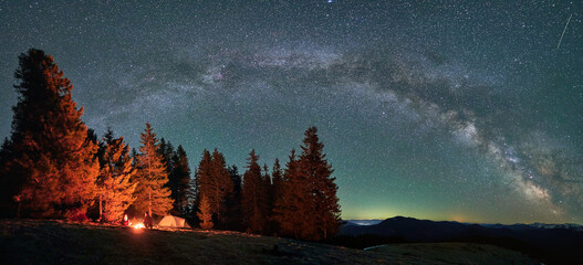 Panorama of starry sky, Milky Way over mountain hills. Silhouettes of two travelers with tent who...