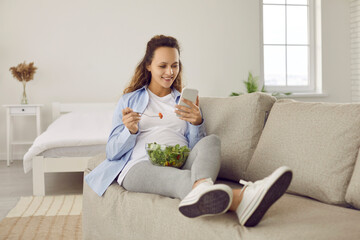 Smiling young pregnant woman relax on couch at home using smartphone having healthy lunch. Happy girl rest on sofa with cellphone eat salad for dinner follow healthy lifestyle. Diet and pregnancy.