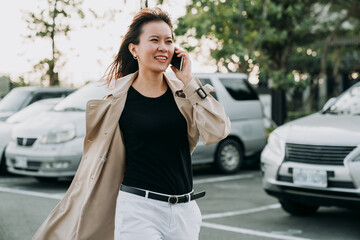 successful asian woman manager in smart casual is walking by a city car parking lot while having a pleasant business talk on the smartphone on a windy day. © PR Image Factory