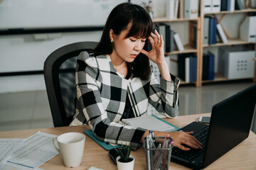 stylish asian female secretary having a sudden headache is rubbing her temple with eyes closed while working on the laptop in the office.