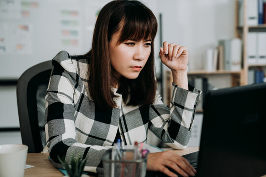 Asian Woman Creative Worker Having Blurred Vision Is Squinting Eyes While Leaning Forward Close To The Computer Monitor Trying To See Clearly At Office Desk.