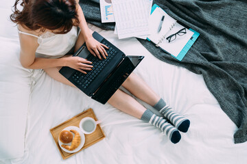 view from top asian woman sitting with extended legs and breakfast is typing financial report on computer in the morning on bed while working from home.