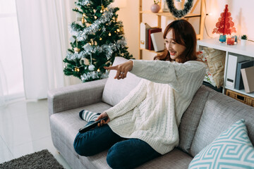 happy asian young lady relaxing on sofa is pointing with finger while laughing at the comedy she’s watching on tv during Christmas vacation days at home.