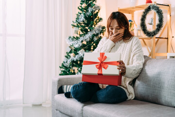 overjoyed asian woman is covering her mouth in surprise for receiving a valuable gift on christmas morning in a festive home living room interior.