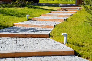 Fototapeta premium The pedestrian path in the park is paved with paving stones, a stone path, stone steps, logs lie on the road, the lawn is mowed, the landscape design of city parks.