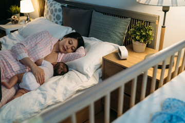 view over the baby crib of loving asian mother wearing pajamas is falling asleep while breastfeeding her newborn kid on bed in a cozy bedroom at night.