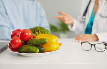 Dietitian or nutritionist giving consultation to patient. Close up of plate of fresh raw vegetables on white table, with doctor and client talking in background. Healthy food, eating healthy concept